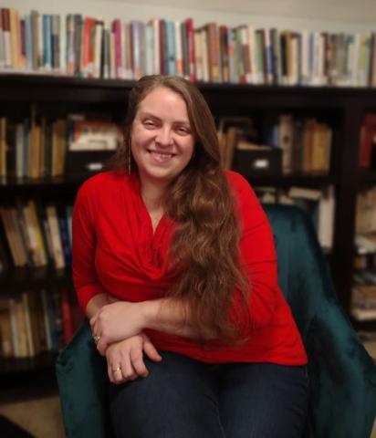 Sarah Wassberg Johnson headshot - a woman with long brown hair wearing a red sweater and sitting in a blue chair in front of a bookcase